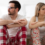 a man and a woman in pajamas sit next to each other, looking away from one another and clearly upset