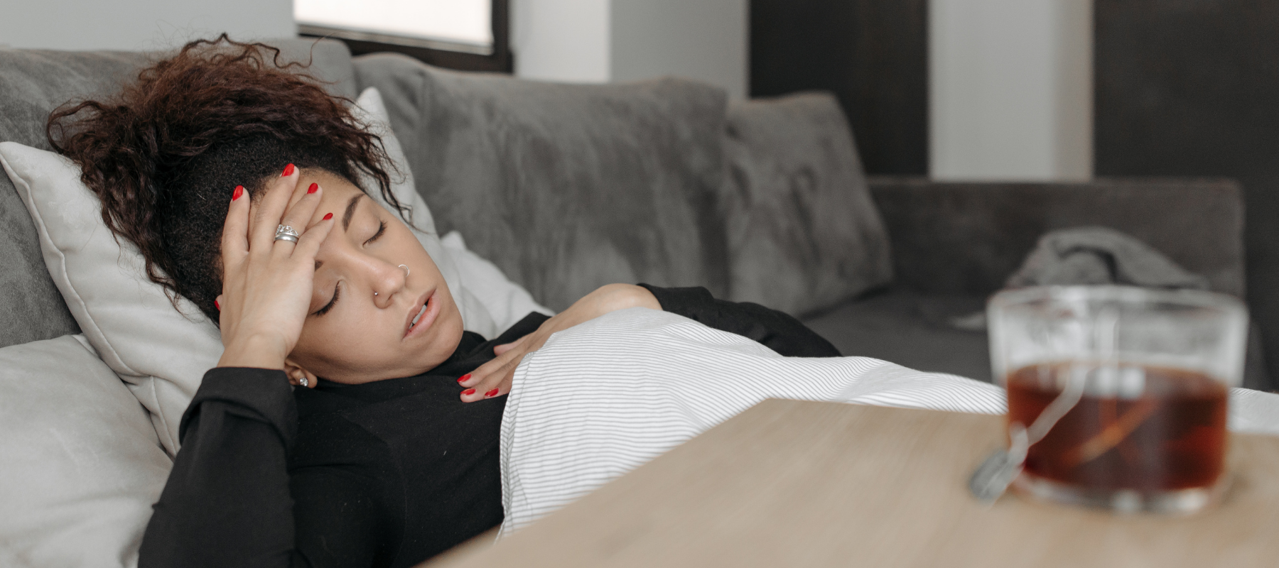 COVID-19 y migraña: Cómo controlar la migraña cuando está enfermo a woman in pain is laying in bed with her hand on her forehead next to a cup of tea on her nightstand